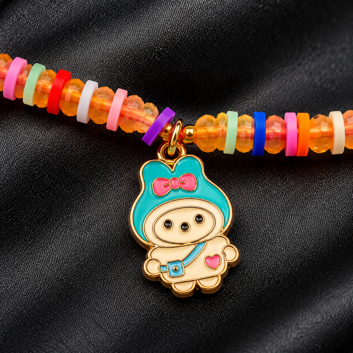 A close-up of a beaded bracelet featuring translucent orange beads and a gold-tone charm of a cute cartoon character with turquoise, pink, and cream enamel details, set against a black fabric background.