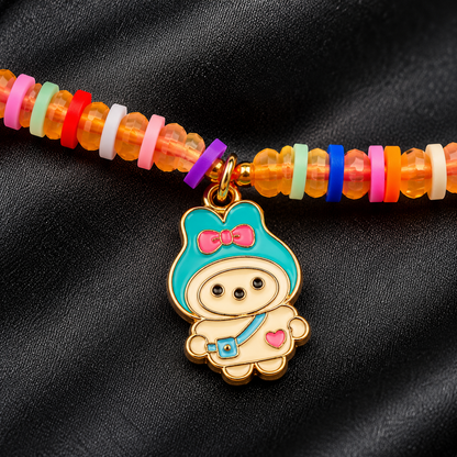 A close-up of a beaded bracelet featuring translucent orange beads and a gold-tone charm of a cute cartoon character with turquoise, pink, and cream enamel details, set against a black fabric background.