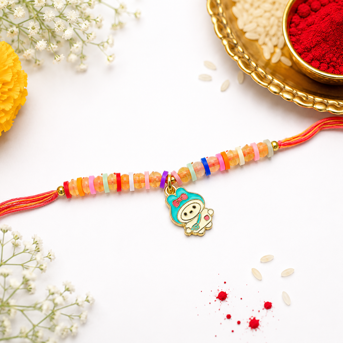 A festive beaded rakhi with a cartoon character charm, displayed on a white background with traditional decorative props including rice, red powder, and baby's breath flowers.