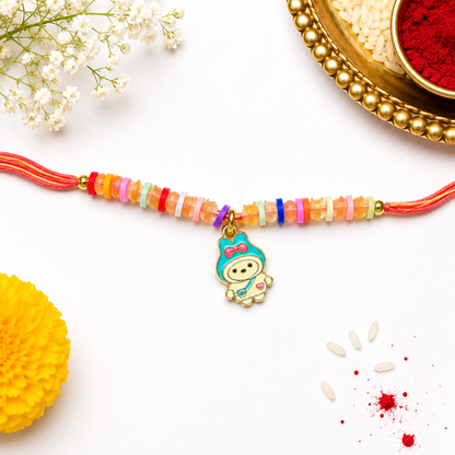 A colorful beaded Rakhi with a cartoon character charm, displayed on a white background with decorative flowers, a yellow flower, and a gold bowl of red powder.