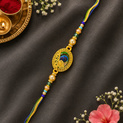A gold-plated peacock-themed Rakhi bracelet with colorful braided cords and pearl-like beads, displayed on a dark satin fabric with decorative floral and diya props.