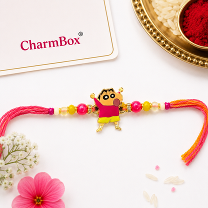 A colorful beaded Rakhi with a Shin-chan character charm, displayed on a white surface with decorative flowers, a gold plate, and a CharmBox card.