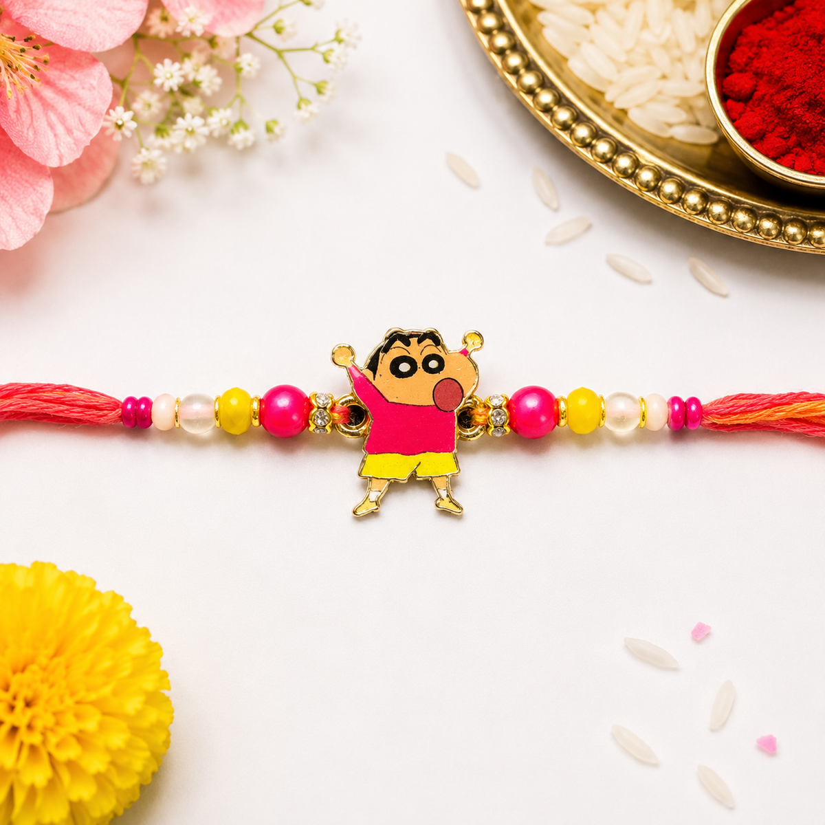 A beaded baby boy's Rakhi featuring a central character charm resembling Shin-chan, styled on a white surface with pink flowers, a yellow pom-pom, and a traditional rice and red powder tray in the background.